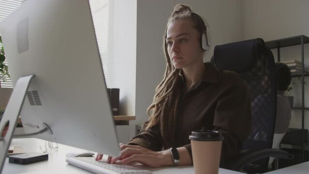 Waist up shot of young Caucasian female programmer with dreadlocks putting on wireless headphones while sitting in front of desktop computer in modern open space office