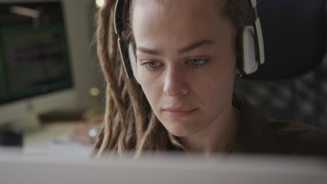 Medium closeup of focused young Caucasian woman with long dreadlocks and wireless headphones program coding on computer in programmers office