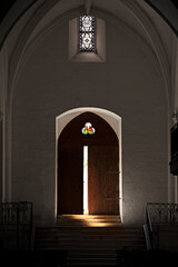 Porte d'entrée d'une église surmontée d'un vitrail en trèfle vue de l'intérieur avec rai de lumière qui éclaire les marches; - obrazy, fototapety, plakaty
