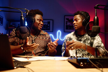 Two young African American men in headphones having discussion of broadcast subject while sitting in studio and talking in microphones