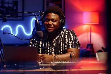 Young African American male host talking in microphone while sitting by workplace and recording new...