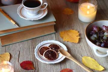 Cup of tea or coffee, plate with desserts, dried oranges, bowl of grapes, scented candles, vintage books, pumpkins and autumn leaves on the table. Autumnal hygge. Selective focus.