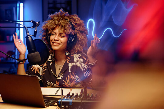 Happy female host in headphones explaining something to online audience while sitting in studio and speaking in microphone