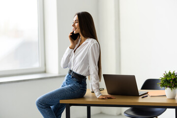 Young woman sales manager talking on cell phone using laptop in office