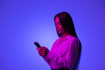 Young woman wearing casual jacket and headphones standing isolated over blue neon light background, using her mobile