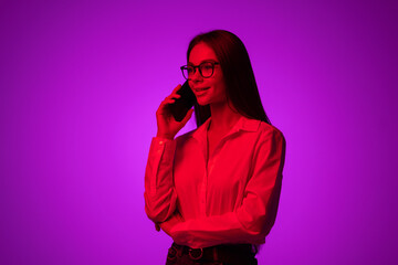 Portrait of young beautiful girl with smartphone isolated on dark background in neon light. Talking on phone.