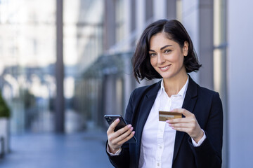 Obraz premium Close-up portrait of a young business woman standing near an office center, holding a phone and a credit card, smiling and looking at the camera