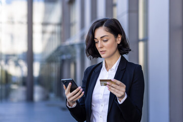 A young business woman is standing on the street near the work office and using a credit card and a phone