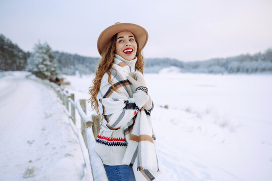 Beautiful young woman with red lips wearing a hat and a light scarf enjoys a winter day in a snowy forest. Happy female tourist posing outdoors. Christmas.