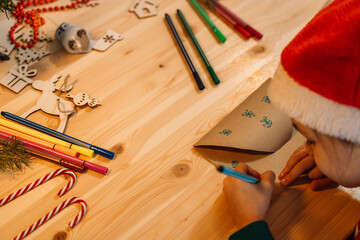 Little girl writes a letter to Santa Claus on a wooden table among pens and Christmas decorations.