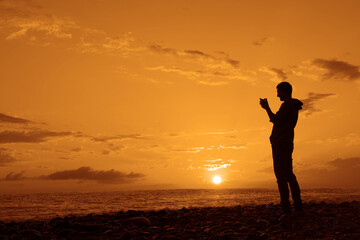 Fototapeta na wymiar black silhouette of Man taking a photo with his mobile phone during the lovely sunset in Black sea. Male traveler make panorama shoot. adult man standing on empty yellow orange beach on background.