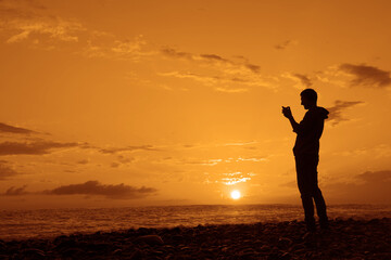 Fototapeta premium black silhouette of Man taking a photo with his mobile phone during the lovely sunset in Black sea. Male traveler make panorama shoot. adult man standing on empty yellow orange beach on background.
