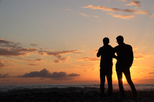 Standing gay two men couple close to each other in front of sea coast background. black silhouette of pair of 2 gays on the beach and sunset. back rear view.