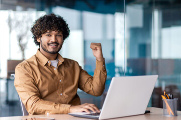 Portrait of winner businessman at workplace, smiling man celebrating successful achievement results and looking at camera, holding hands up victoriously.