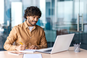 Man studying online remotely, businessman improving qualifications at the workplace inside office, writing down data in a notebook, watching a video course, satisfied with high results, smiling.