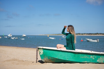 Young, beautiful, blonde, green-eyed, young woman in a green dress, sitting in a boat, covering herself from the sun with her hand, relaxed and calm, on the beach, with the sea in the background.
