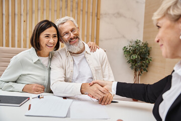 middle aged happy man shaking hands with realtor near wife and making deal in real estate office