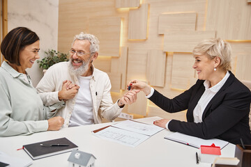 middle aged realtor giving key from new house to happy married couple in real estate office