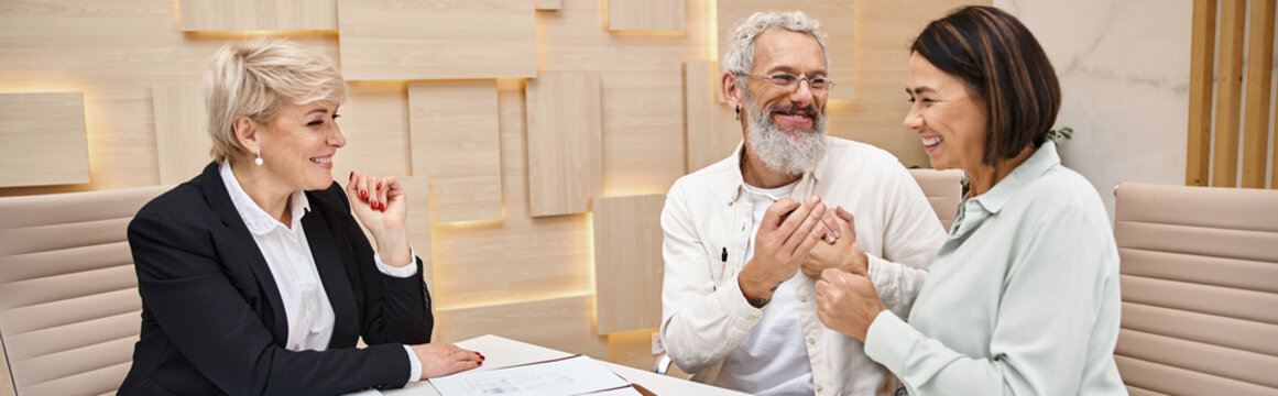 Middle Aged Realtor Looking At Happy Married Couple Holding Key From New House In Office, Banner