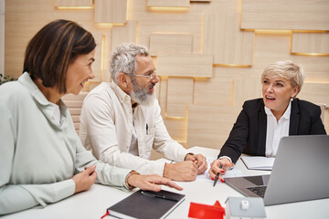 Fototapeta premium happy middle aged married couple discussing project of new home with realtor in real estate office