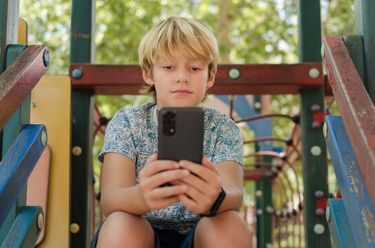 10 years old cute blonde boy using smartphone outdoors. Portrait of concentrated kid sitting in a playground alone holding mobile phone looking social media. Rest, education and technology concept.