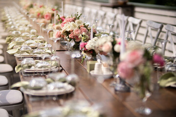 A large, long, decorated, wooden table and chairs, covered with a white tablecloth with dishes, flowers, candles.
