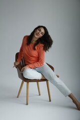Beautiful woman of 30 years old sitting on chair, white background. Portrait of brunette girl smile. Orange sweater and light jeans. Woman barefeet, thoughtful.