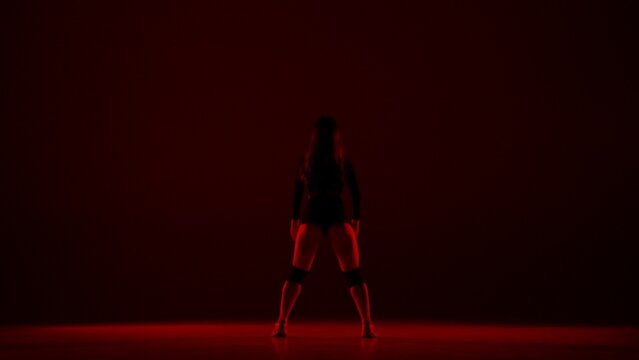 In the frame on a dark background, a young woman, on it shines a red light stands a woman. Demonstrates a dance movement in the style of twerk. She is sexy, rhythmic. She is wearing open clothes.