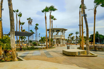 Panoramic view of promenade or malecon with its kiosk, palm trees, concrete benches, sky covered...