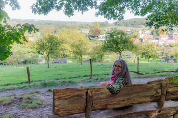 Senior adult woman sitting on wooden bench, turned away looking at camera, cheerful expression, Dutch village and countryside in misty background, sunny day in Sweikhuizen, South Limburg, Netherlands