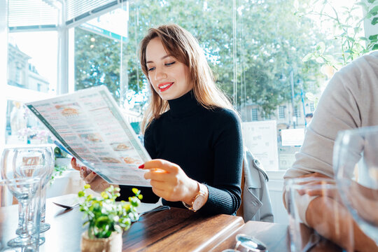 Young woman reading menu sitting with friend at casual cafe. Smiling woman checking menu, choosing meal or drink, making order. Try new foods while traveling. real people. copy space. Selective focus.