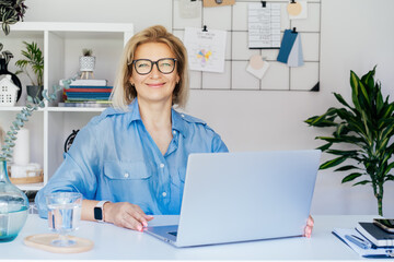 Smiling mature businesswoman with laptop at her office workplace, confident middle-aged experienced...