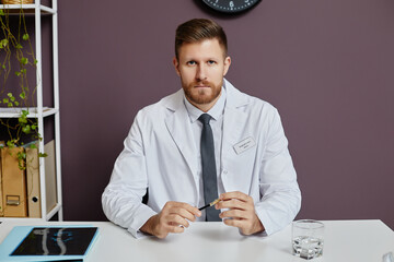 Front view portrait of professional bearded doctor sitting at workplace in clinic and looking at camera with serious face expression