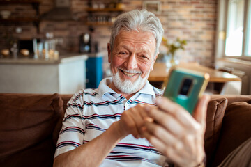 Smiling elderly man sitting on couch at home using mobile phone