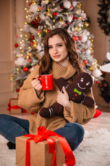 Beautiful young woman with a red mug of coffee in her hands against the background of a Christmas tree.