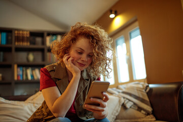 Young smiling redhead woman using a smart phone while sitting on a couch in the living room - obrazy, fototapety, plakaty