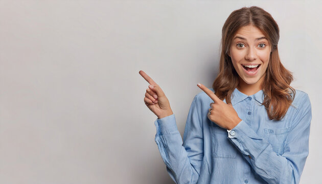 horizontal shot of pretty surprised cheerful young woman pointing to empty copy space advertises product or tells about awesome offer dressed in casual clothing isolated over white background