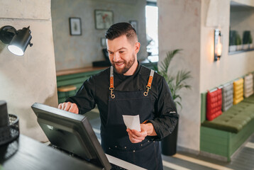 Stylish bearded smiling waiter dressed black uniform processing customer orders using point of sale...
