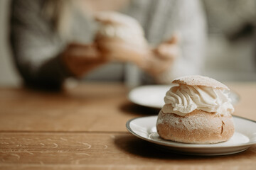 Focus on traditional Swedish pastry, semla, on a plate at wooden table. In the background, out of focus, a person is about to eat pastry. Photo taken in Sweden.