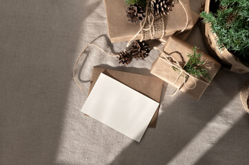 Blank paper card, envelope mockup, crafted Christmas gift boxes on linen neutral beige tablecloth...