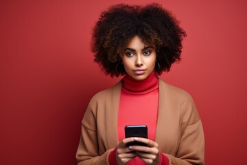 Fototapeta na wymiar Studio portrait of beautiful African American woman with smartphone in red clothes against red background. Positive girl with Afro haircut texting message, enjoying online communication, using app.