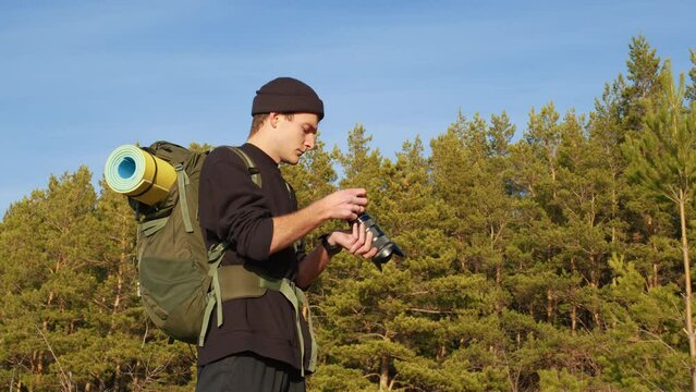 Handsome guy with professional camera takes pictures in woods, evaluates result. Male tourist with backpack, mat is shooting of forest landscapes. Cameraman working on nature. Side view