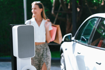 Young woman travel with EV electric car charging in green sustainable city outdoor garden in...