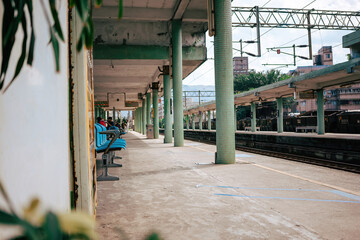 An almost deserted train platform stretches out, characterized by teal benches and weathered...