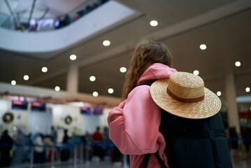 Back view of female traveler with straw hat and backpack awaits flight in airport terminal for...