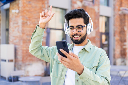 A young man in headphones is walking around the city, joyful, smiling, listening to music online, using an application on the phone