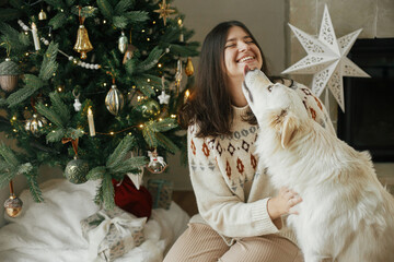 Merry Christmas! Happy woman in cozy sweater hugging and playing with cute white dog on background...