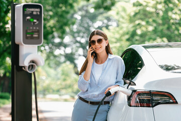 Young woman talking on smartphone while recharging electric car battery charging from EV charging...
