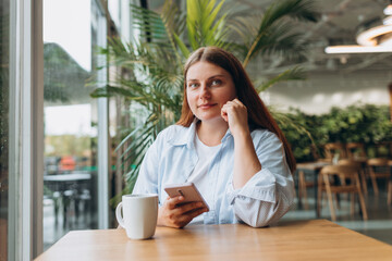 Young happy businesswoman smiling and using smart phone in a cafe. Person sitting at table and using smartphone indoors. Online education, order, working or shopping concept
