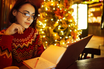 Young smart student immersed in her studies with laptop in cozy Christmas cafe on the eve winter holidays - 679382163